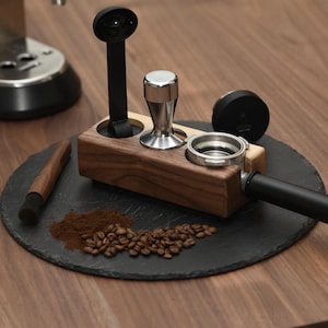Könnte beinhalten: Ein Kaffee-Station-Setup mit einem Holzhalter mit einem Tamper, einem Siebträger und einem Löffel. Ein schwarzer Schieferuntersetzer hält Kaffeepulver und -bohnen, mit einer Bürste und einem schwarzen Deckel. Eine Kaffeemaschine befindet sich im Hintergrund.