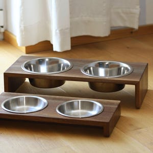 Könnte beinhalten: Zwei Futternäpfe aus Holz für Haustiere mit Edelstahleinsätzen. Die Schalen stehen auf einem dunkelbraunen Holzsockel. Ein Schalenständer hat zwei Schalen, der andere eine Schale. Die Schalen stehen auf einem Hartholzboden.