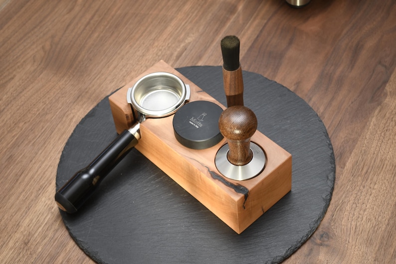Op de afbeelding: Een houten espressostation met een portafilter, een tamper en een zwart puckscherm. Het station is gemaakt van lichtgekleurd hout en staat op een donkergrijze leistenen onderzetter. De tamper heeft een houten handvat en een roestvrijstalen basis.