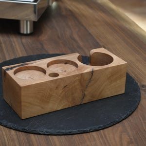 Op de afbeelding: Een lichtbruin houten espressostation met drie cirkelvormige uitsparingen en een gebogen houder. Het station staat op een zwart, rond oppervlak. De houtnerf is zichtbaar.
