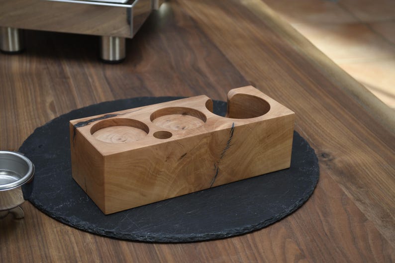 Op de afbeelding: Een lichtbruin houten espressostation met drie cirkelvormige uitsparingen en een gebogen houder. Het station staat op een zwart, rond oppervlak. De houtnerf is zichtbaar.