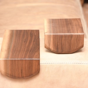 Könnte beinhalten: Zwei Holzarmlehnenbezüge mit einer satten, dunkelbraunen Maserung. Die Bezüge haben eine gebogene Vorderkante und eine flache Oberseite, die für die Armlehnen eines Sofas konzipiert sind. Die Holzmaserung ist sichtbar und verleiht dem Möbel eine natürliche Ästhetik.