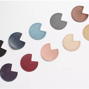 Op de afbeelding: Een verzameling kleurrijke, halfronde leren stukken in verschillende tinten, waaronder zwart, marineblauw, blauw, rood, roze en beige. Elk stuk heeft een gestikte rand en een uitgesneden gedeelte, waardoor een unieke vorm ontstaat. De stukken zijn op een witte ondergrond geplaatst.