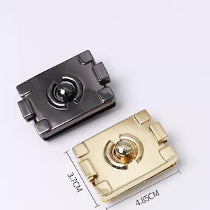 May include: Two metal purse clasps. One is a dark gunmetal color, the other is gold. The gold clasp measures 1.9 inches by 1.5 inches. Each clasp has a central circular element and four square corner accents.