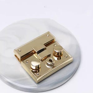 May include: A gold-colored metal purse lock with a rectangular base and two round knobs. The lock is set on a gray and white marbled surface. The lock has a rectangular clasp and small screw holes.