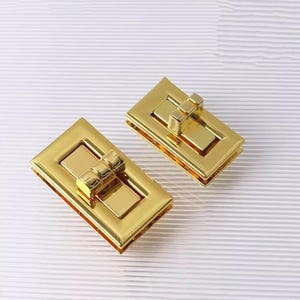 May include: Two gold-colored rectangular purse clasps. Each clasp features a rectangular frame with a central bar and a smaller, decorative bar. The clasps are made of shiny metal and are set against a white, textured background.