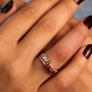 May include: A rose gold ring with a row of small, vibrant red gemstones and a second ring with clear stones. The ring is on a finger with dark red nail polish. The background is a denim top.