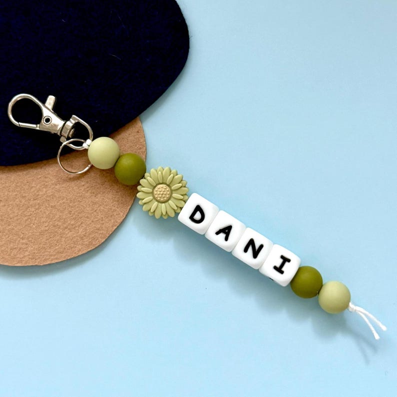 Puede incluir: Un llavero personalizado con el nombre "DANI" escrito con cuentas de letras blancas. El llavero presenta cuentas verdes, un dije en forma de margarita y un cierre plateado. El fondo es azul claro.