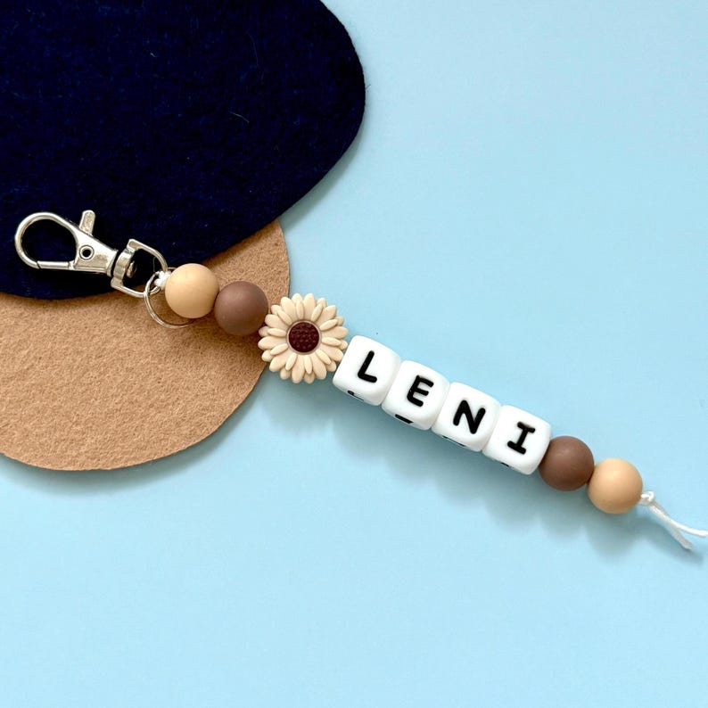 Puede incluir: Un llavero personalizado con el nombre "LENI" escrito con cuentas de letras blancas. El llavero presenta un cierre plateado, cuentas beige y marrones, y una peque&ntilde;a flor de margarita. El fondo es azul claro.