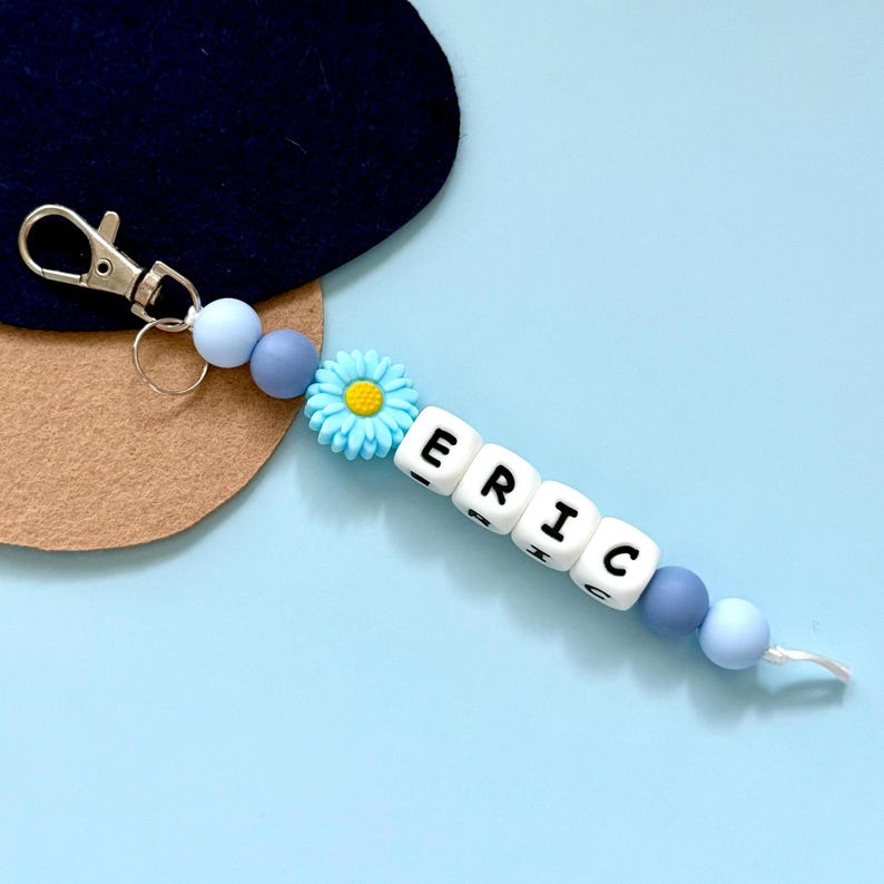 Puede incluir: Un llavero personalizado con el nombre "ERIC" escrito con cuentas de letras blancas. El llavero presenta cuentas azules y grises, un dije de margarita azul y un cierre plateado. El fondo es azul claro.