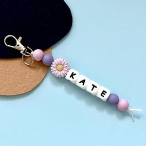 Puede incluir: Un llavero con el nombre "KATE" escrito con cuentas blancas. El llavero presenta cuentas rosas y moradas, un dije de margarita y un cierre plateado. El fondo es azul claro.