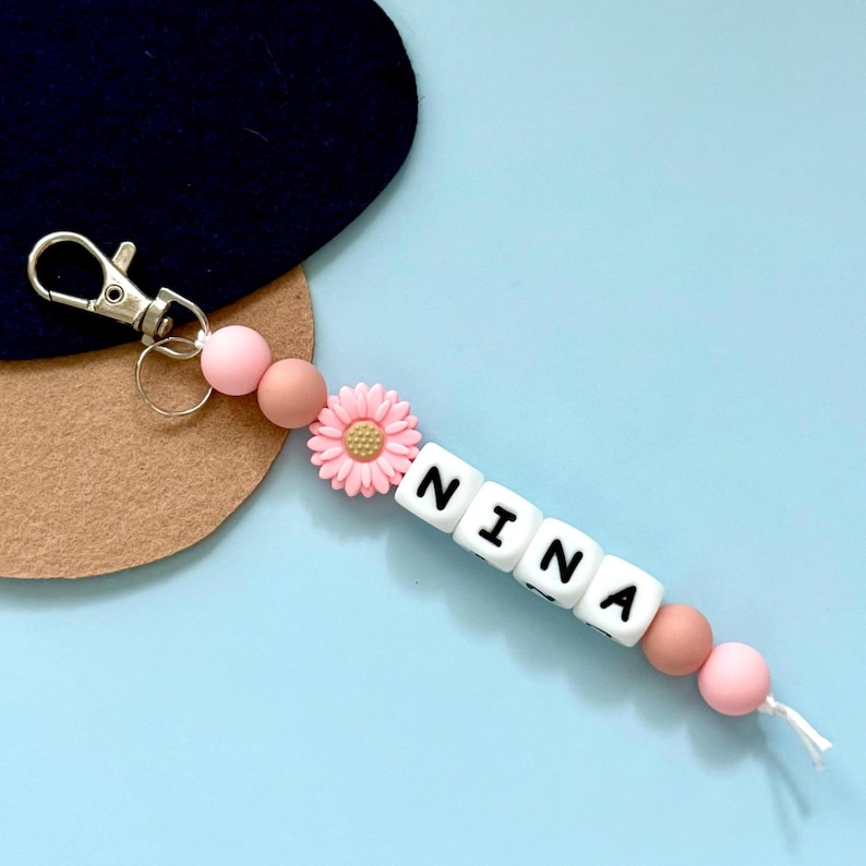 Puede incluir: Un llavero personalizado con el nombre "NINA" escrito con cuentas de letras blancas. El llavero presenta cuentas rosas y marrones, una flor de margarita rosa y un cierre plateado. El fondo es azul claro.