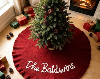 Könnte beinhalten: Ein kreisförmiger, dunkelroter gestrickter Weihnachtsbaumteppich mit gerüschtem Rand und dem weißen gestickten Text "The Baldwins" in der Mitte. Der Baumteppich liegt um den Fuß eines grünen Tannenbaums.