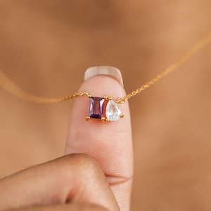 May include: A delicate gold necklace featuring two gemstone pendants. One pendant is a rectangular purple stone, and the other is a pear-shaped clear stone. The necklace is held up by a finger.
