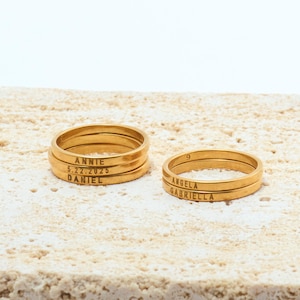 May include: Gold-coloured stackable rings with engraved names and dates. One stack includes the names "ANNIE", "DANIEL", and the date "5.22.2025". The other stack includes the names "ANGELA" and "GABRIELLA". The rings are displayed on a textured, light-coloured surface.