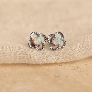 May include: A pair of silver-toned stud earrings. Each earring features a four-petal flower design with a textured border. The center of each flower holds an opal stone with iridescent colors. The earrings are displayed on a beige fabric.