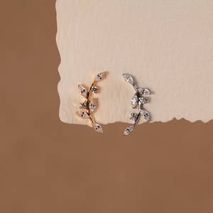 May include: Two ear climbers shaped like curved branches with leaves. One climber is gold-toned with clear marquise and round-cut stones, and the other is silver-toned with clear marquise and round-cut stones, both featuring a delicate vine design.