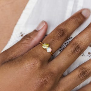 May include: A gold ring featuring two gemstones: a pear-shaped yellow stone and a round, iridescent opal. The ring is set on a slender gold band, creating a delicate and elegant design. The ring is shown on a person's finger.
