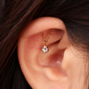May include: A gold-toned ear piercing jewelry piece featuring a delicate chain that connects two points of the ear cartilage, with a single clear, pear-shaped, teardrop-style gemstone charm suspended from the center of the chain.