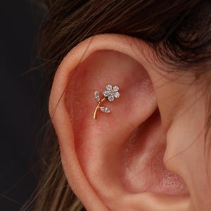 May include: A gold-toned ear piercing stud shaped like a flower with a stem and two leaves. The flower head is composed of six small, clear, round-cut stones, while the stem and leaves are accented with two clear, marquise-cut stones.