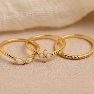 May include: Three gold rings with opal detailing. One ring has a chevron design with opal stones, another features a floral arrangement of opals, and the third has a textured band. The rings are displayed on a neutral background.