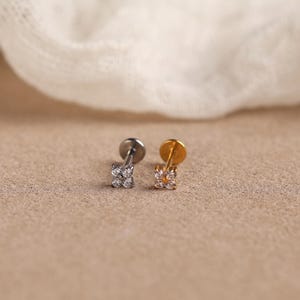 May include: Two flat-back stud earrings, one in silver tone and one in gold tone, each featuring a small cluster of four clear, round-cut stones arranged in a square shape. The earrings have a straight post and a circular flat backing disc.