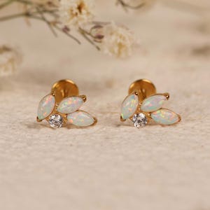 May include: A pair of gold-tone stud earrings featuring three marquise-cut white opals arranged in a leaf-like cluster, accented by a single small round clear gemstone at the base of each earring, with a flat circular backing visible on each piece.
