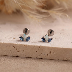 May include: A pair of stud earrings featuring a central marquise-cut white opal flanked by two smaller blue marquise-cut gemstones on each side. The earrings have a metallic post back and are designed with a curved, multi-stone arrangement.