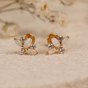 May include: A pair of gold stud earrings featuring a cluster design. Each earring consists of three marquise-cut white opals and two round-cut clear stones arranged in a floral-like pattern. The earrings have a flat gold backing post.