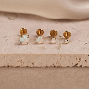 May include: Four gold-tone labret studs featuring round, iridescent white opal stones in prong settings. The studs are arranged in a row, decreasing in size from left to right, each with a flat circular back and a thin metal post for piercing.