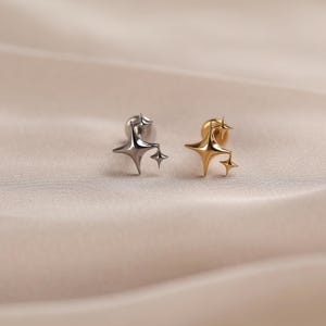 Puede incluir: Dos pendientes de botón con un diseño de estrella de cuatro puntas con una estrella más pequeña adjunta. Un pendiente tiene un acabado en tono plateado pulido, mientras que el otro tiene un acabado en tono dorado pulido. Ambos tienen cierre plano.