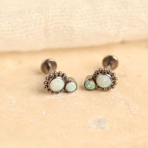 May include: Two silver-toned stud earrings with a cluster of small beads around a large, round, white opal stone and a smaller opal stone. The earrings are displayed on a light beige surface.