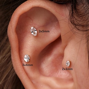 Puede incluir: Tres pendientes de botón de forma ovalada con piedras transparentes engastadas en metal dorado. Los pendientes están etiquetados con los tamaños 3x5mm, 3x4mm y 2x3mm, indicando sus dimensiones respectivas sobre el cartílago y el lóbulo.