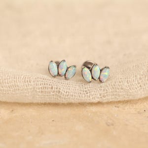 May include: A pair of stud earrings featuring three marquise-cut white opals arranged in a fan-like cluster. The stones are set in a metallic base with a dark finish, showcasing iridescent flashes of color within each individual opal gemstone.