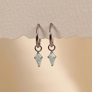 Può includere: Un paio di orecchini a cerchio color argento con piccoli ciondoli in opale a forma di aquilone. Ogni orecchino è composto da un sottile cerchio con una pietra di opale bianco sfaccettata che pende, mostrando riflessi iridescenti.