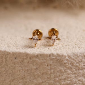 May include: A pair of gold-toned stud earrings featuring a four-pointed star design with a central pear-shaped clear gemstone. Each earring has a flat circular backing and a metallic finish, showcasing a symmetrical starburst shape with sharp points.