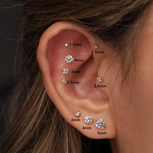 May include: A collection of round, clear, prong-set stud earrings in various sizes, including 1.5mm, 2.0mm, 2.5mm, 3mm, 4mm, and 5mm, arranged along the cartilage and lobe, with each size clearly labeled with text next to the corresponding earring.