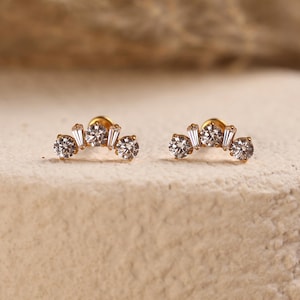 May include: A pair of gold stud earrings, each featuring a curved design set with three round brilliant-cut clear stones and two smaller baguette-cut clear stones arranged in an alternating pattern along the metal arc.