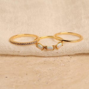 May include: Three gold rings are displayed on a cream-colored cloth. One ring is adorned with small, light-colored stones, another features a row of white opal-like stones, and the third is a simple band. The rings are set against a neutral background.