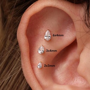 May include: Three pear-shaped clear stud earrings arranged vertically in an ear. The top stud is 6x4mm, the middle stud is 3x4mm, and the bottom stud is 2x3mm. Each stud features a faceted stone set in a metallic prong setting, creating a clear, sparkling appearance.