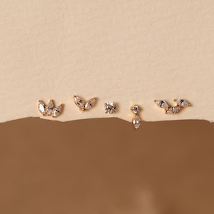 May include: Five gold stud earrings featuring clear, faceted stones in various arrangements, including marquise and round cuts. The set includes cluster designs, a single round stone, and a drop-style earring with a marquise stone.