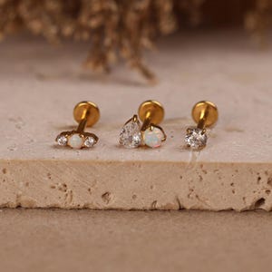 May include: Three gold-tone stud earrings with flat back posts. The left earring features a central opal flanked by two clear stones. The middle earring has a pear-shaped clear stone and a round opal. The right earring is a single round clear stone.