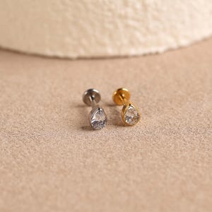 Puede incluir: Dos pendientes de botón con parte posterior plana y una piedra transparente en forma de lágrima. Uno tiene un poste y engaste de metal plateado, mientras que el otro tiene un poste y engaste de metal dorado. Ambos tienen una base circular plana.