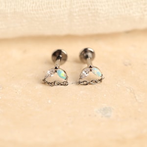 May include: A pair of dark metal flat back stud earrings featuring a whale tail design. Each earring is adorned with one clear marquise-cut stone and one iridescent opal-like marquise-cut stone, connected by a delicate dark metal chain link.