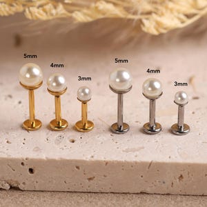 Peut inclure: Un ensemble de six boucles d'oreilles labret avec des perles sphériques blanches montées sur des tiges métalliques. Le lot comprend trois tiges dorées et trois argentées, marquées des tailles 3mm, 4mm et 5mm pour le diamètre des perles.