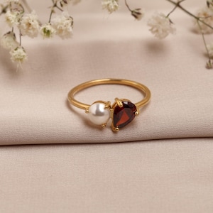 May include: A gold ring featuring a white pearl and a red gemstone. The ring is set on a beige fabric, with white flowers in the background. The ring's design is elegant and eye-catching, suitable for special occasions.