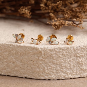 May include: Four gold-tone stud earrings featuring white opal-like marquise stones and small clear round crystals. Each earring has a flat back post design. The pieces are arranged in a row, showcasing leaf-like and floral-inspired arrangements.