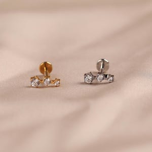 May include: Two flat back labret studs, one in gold tone and one in silver tone, each featuring a horizontal bar set with three clear round stones of varying sizes arranged in a cluster design for body piercing jewelry.