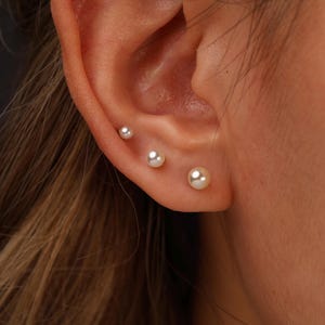 May include: Three white spherical pearl stud earrings arranged in a vertical line along the earlobe and cartilage. Each earring features a smooth, lustrous white pearl set on a small metal post, creating a graduated size effect from smallest to largest.