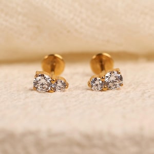 May include: A pair of gold-tone stud earrings, each featuring two round, clear, faceted stones set side-by-side in a metal base. The earrings have a flat, circular backing post, designed for secure placement, showcasing a polished metallic finish.